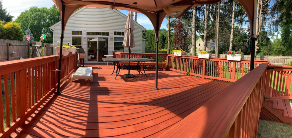 A spacious wood deck with a rich red stain, outdoor furniture, and a pergola overlooking a backyard.
