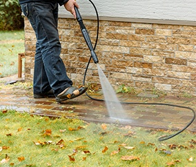A worker power washing a paver walkway next to a stone facade, clearing away dirt and fallen leaves.